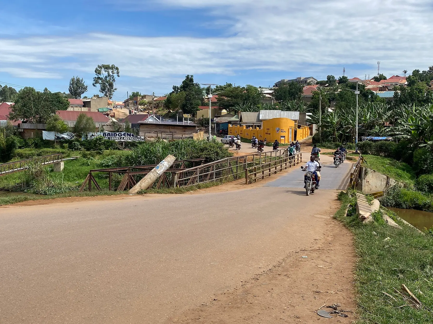 The road across the Katete bridge in Mbarara, with motorcycles crossing and shopfronts and houses on the far bank